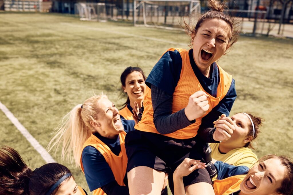 Cheerful team of female soccer players celebrating victory and carrying on of teammates who is shouting out of joy on stadium